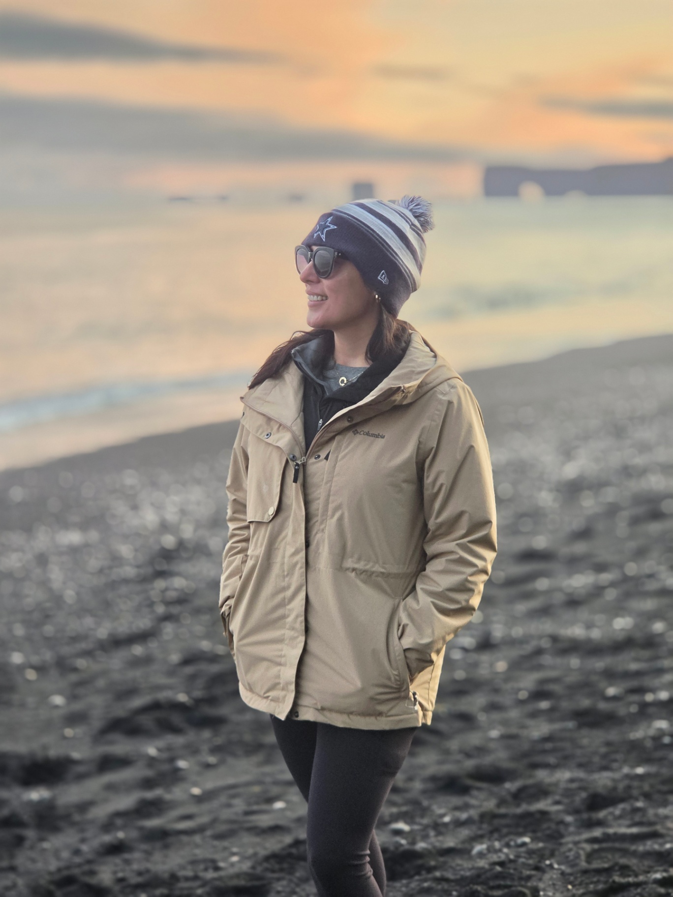 Woman in a tan jacket on a black sand beach at sunset